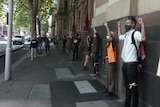Masked people line up at regular intervals along with the wall of a state building with their fists in the air.
