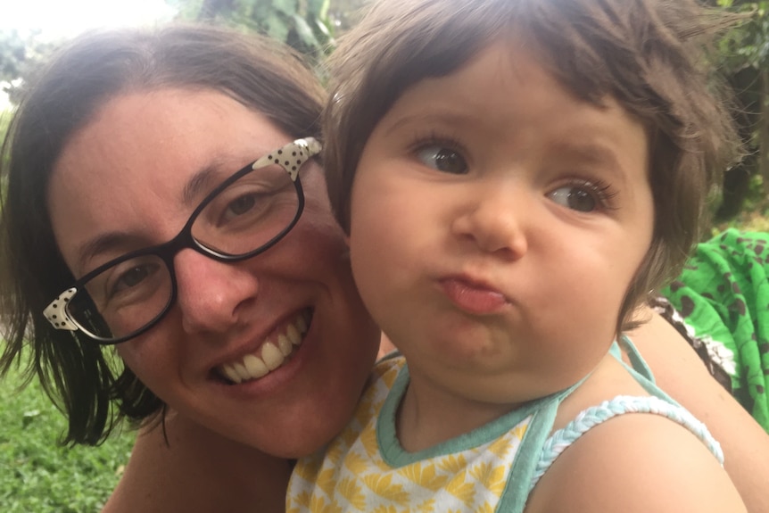 A smiling woman with brown hair and glasses hugs a toddler puffing her cheeks out playfully.