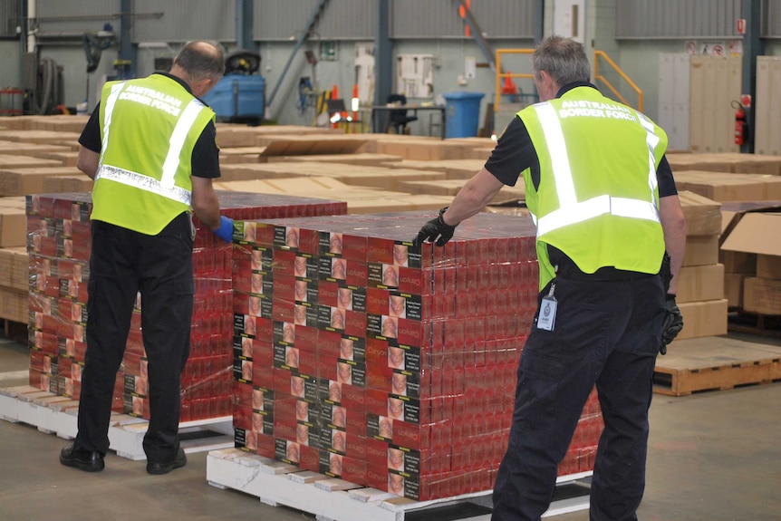 Two Australian Border Force officers stand alongside two pallets stacked with red boxes of illicit cigarettes.