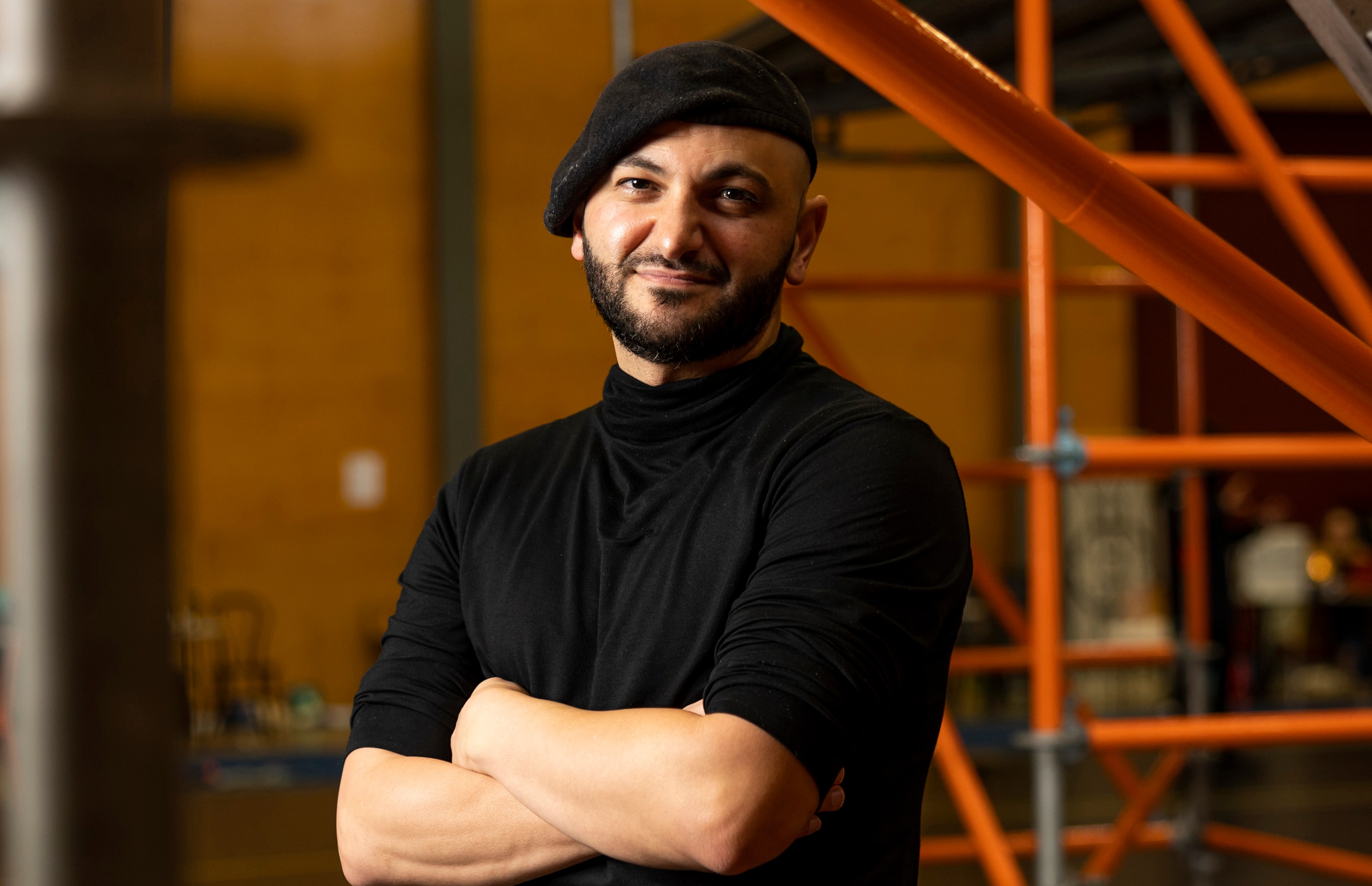 A 30-something Lebanese Australian man wearing a beret stands with his arms folded, amongst scaffolding in a rehearsal space