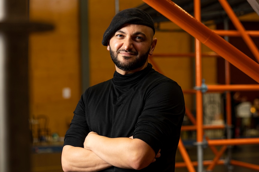 A 30-something Lebanese Australian man wearing a beret stands with his arms folded, amongst scaffolding in a rehearsal space