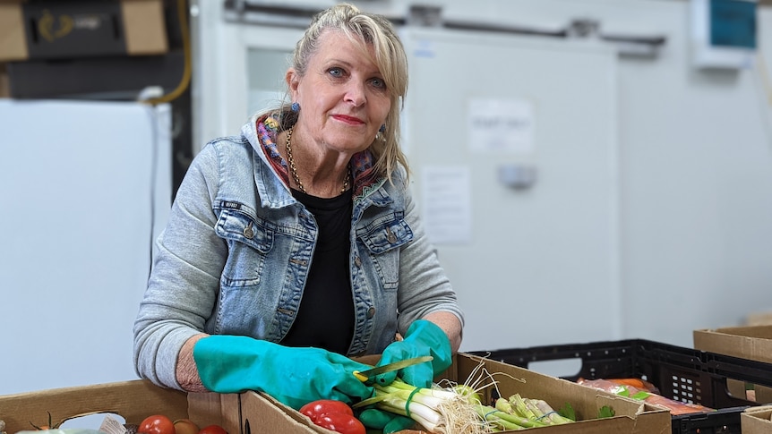 Bev leans over boxes of fresh vegetables wearing green gloves with coolroom behind her