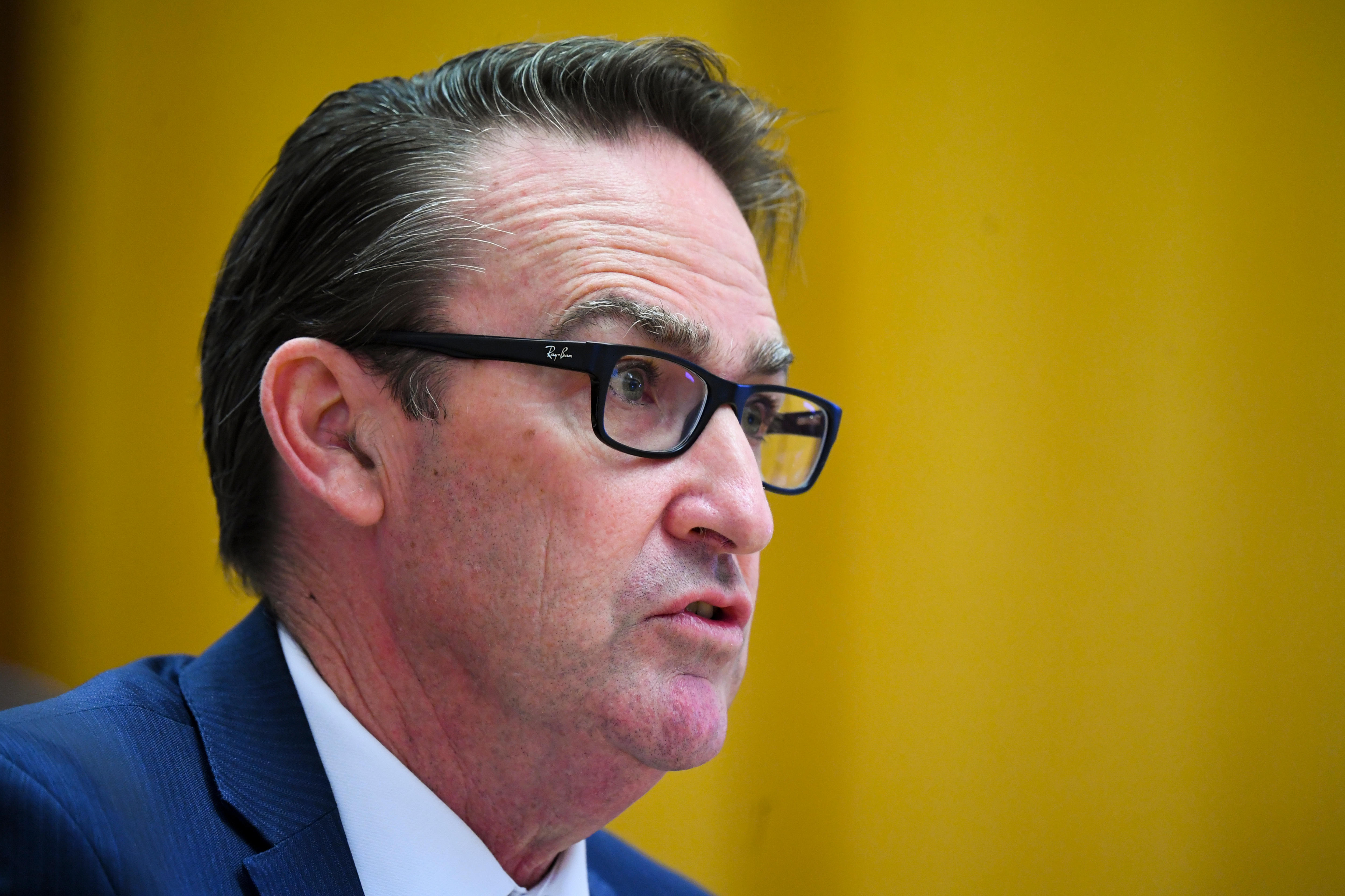 A side-on close shot of a man wearing a blue suit and black glasses.
