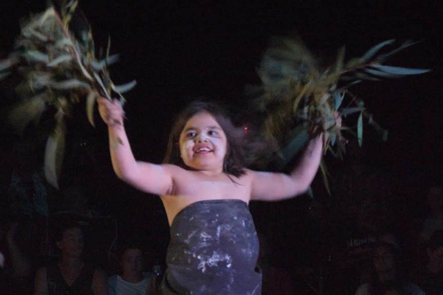 An Aboriginal girl in a black wrap holds up branches of gum leaves.