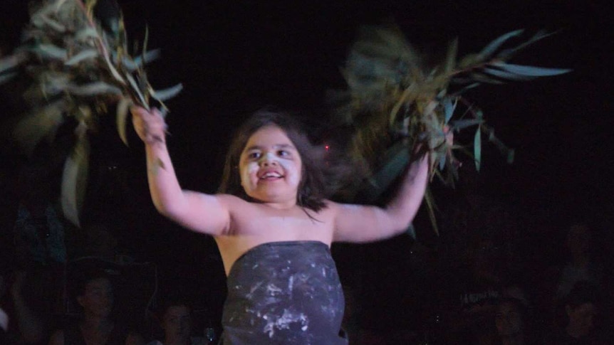 An Aboriginal girl in a black wrap holds up branches of gum leaves.