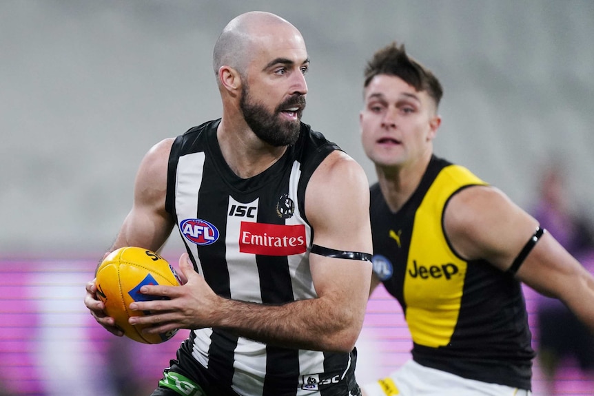A Collingwood AFL player looks to his left as he runs with the ball in both hands in front of a Richmond opponent.