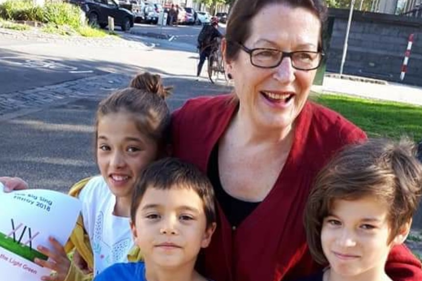 A woman with her arms around three children who are all standing on grass outside.