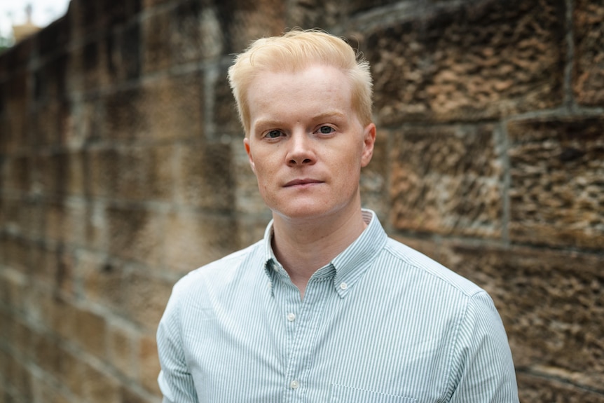 A man looks at the camera with a neutral expression. He is standing in front of a brick wall.