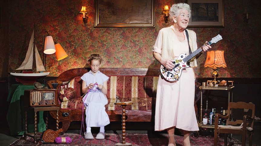 An older woman stands up playing a guitar while a young girl sits on a lounge and knits