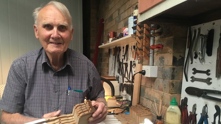 Older man stands in workshop holding wooden block to make piano keys.