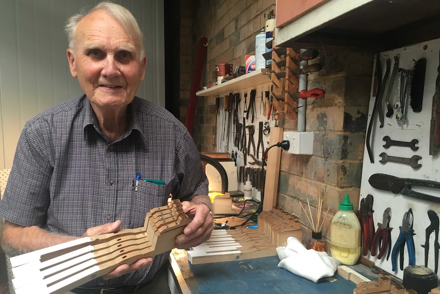 Older man stands in workshop holding wooden block to make piano keys.