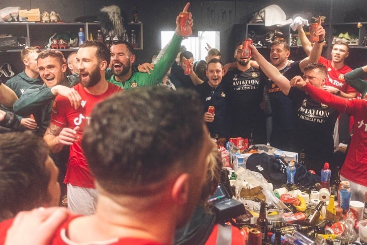 A group of soccer players in red, black and green shirts hold cans of beer and cheers in celebration.