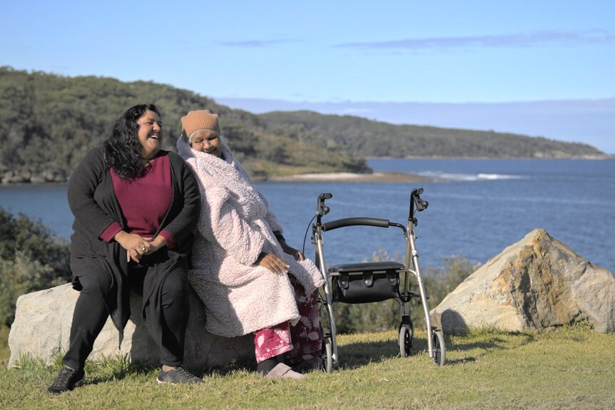 An elderly woman with a walker and a younger woman sit together on a rock overlooking the sea, laughing together.
