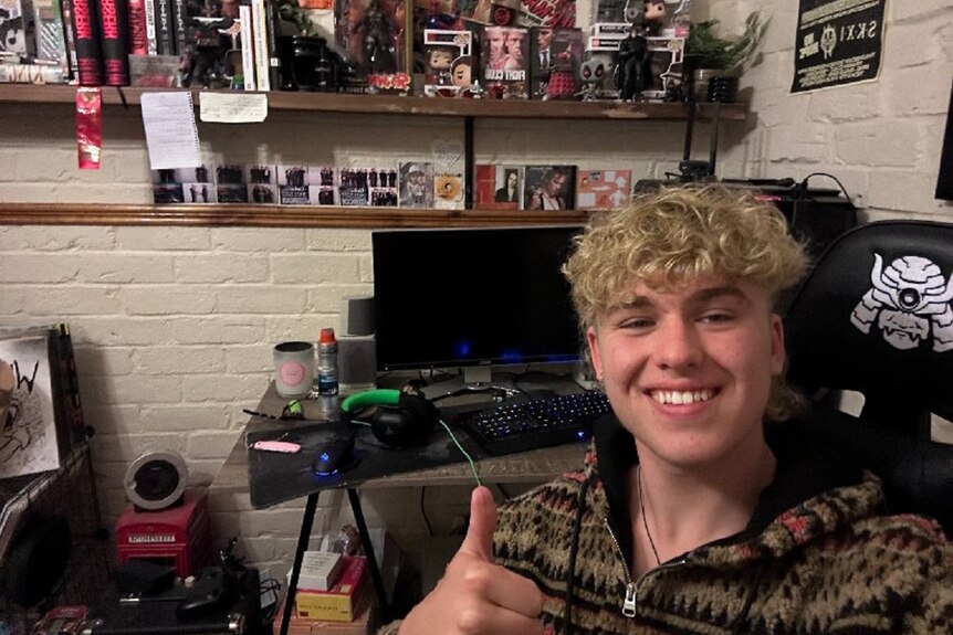 A blonde teenage boy sitting in front of a computer in a gaming chair giving a thumbs up.