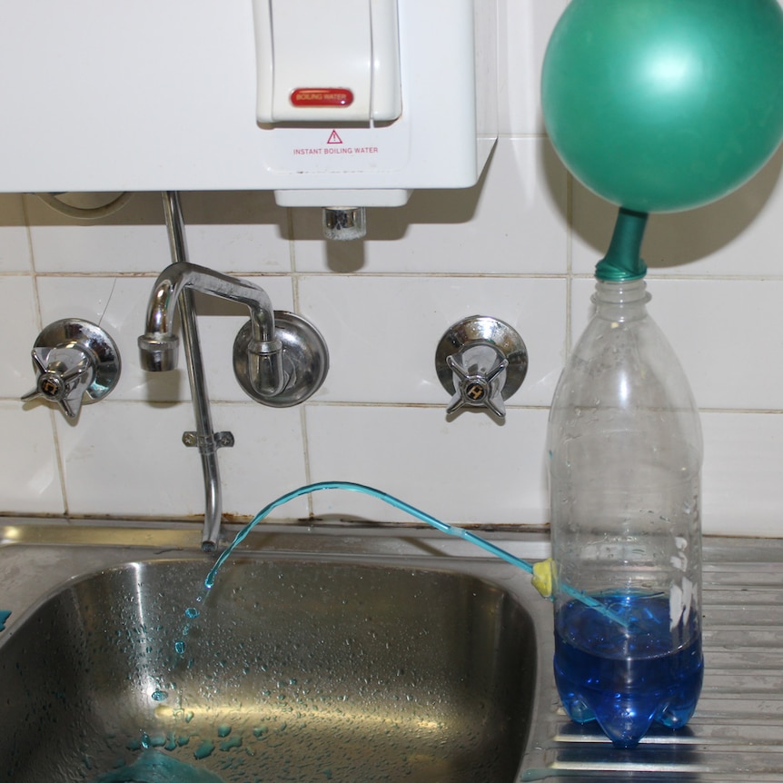An inflated balloon is attached to the top of a 2-litre bottle. Blue water is being pushed out its side through a straw.