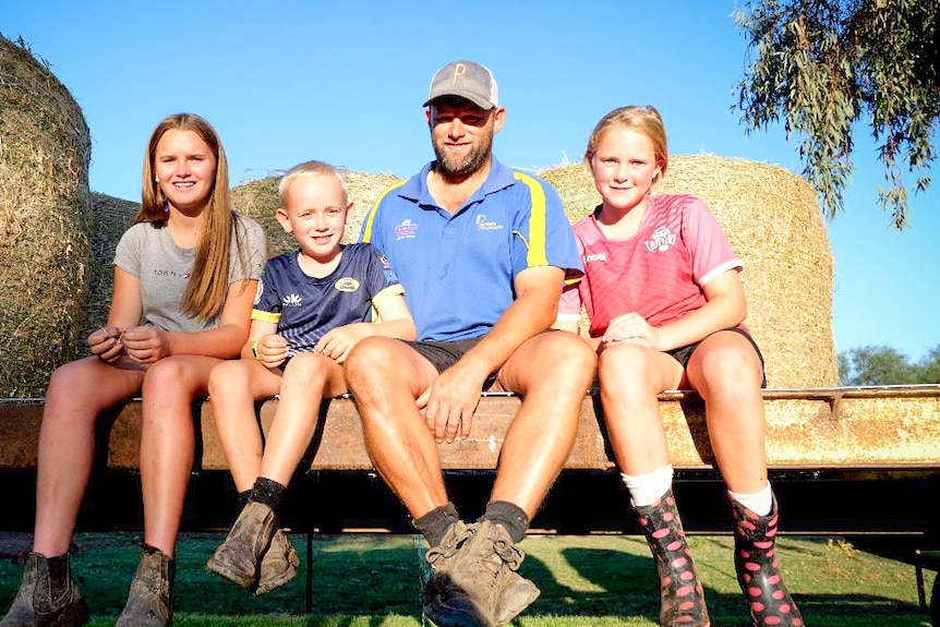 Three children and a man sitting on a low bench in front of hay bales.