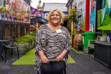 A woman sitting on a chair in a colourfully painted outdoor alleyway smiles for a photo.