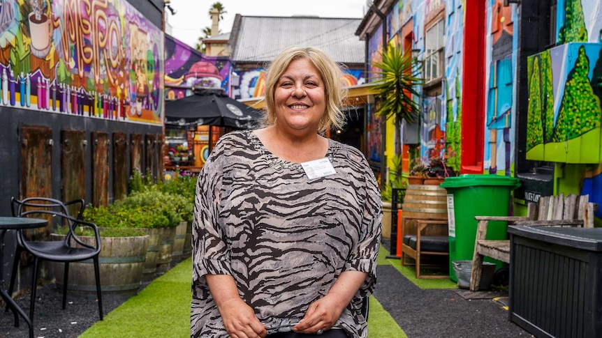 A woman sitting on a chair in a colourfully painted outdoor alleyway smiles for a photo.