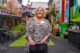 A woman sitting on a chair in a colourfully painted outdoor alleyway smiles for a photo.