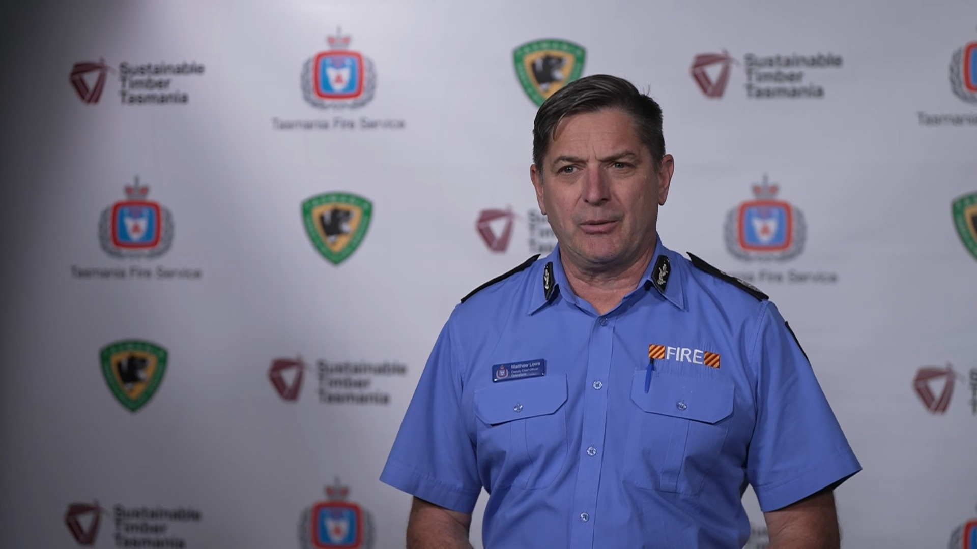 Matt Lowe stands infront of a background with logos of the TFS, PWS and Sustainable Timber Tasmania