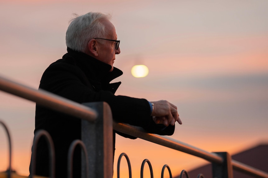 A man with grey hair and glasses stands on a waterfront as the sunsets