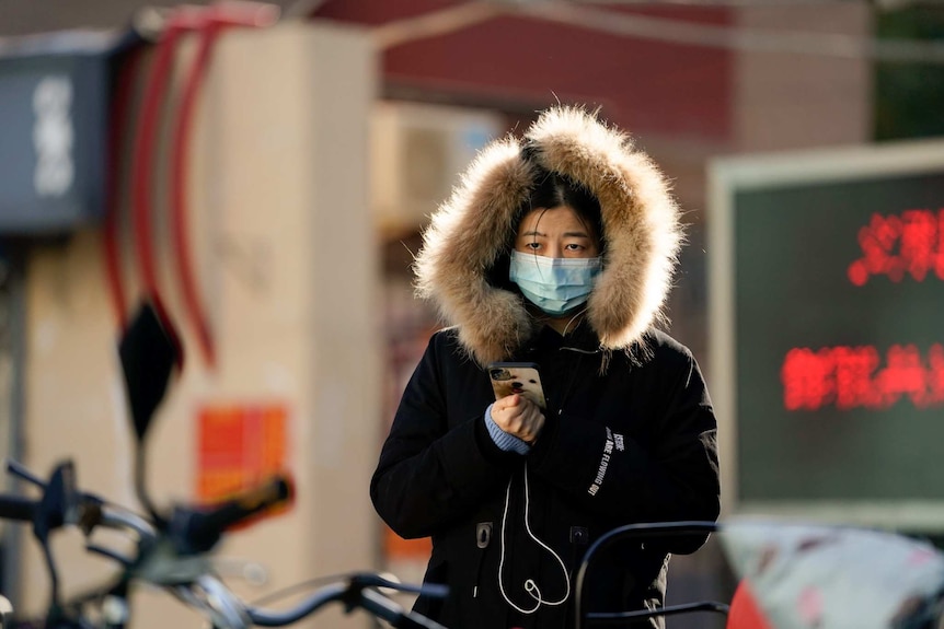 A woman wearing a mask and a fur lined coat holds her phone as she walks holding her phone.