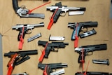 More than a dozen handguns lie on a table with red tags on them.