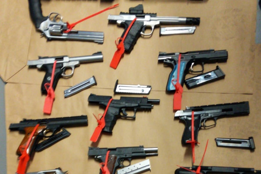 More than a dozen handguns lie on a table with red tags on them.