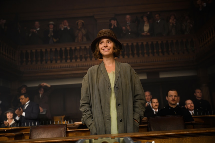A woman in 1920s costume stands in the dock of a crowded courtroom, smiling cheekily with ther hands in her pockets.