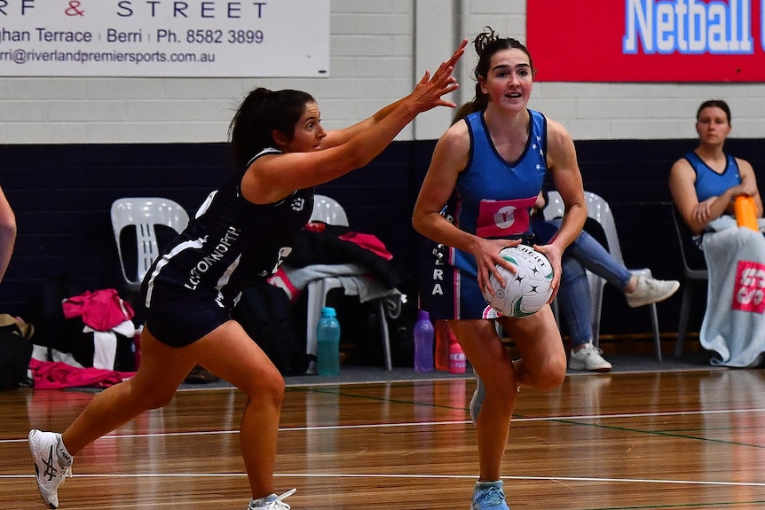 A woman wearing a blue netball uniform holding a netball while a woman wearing a navy uniform tries to block a pass. 