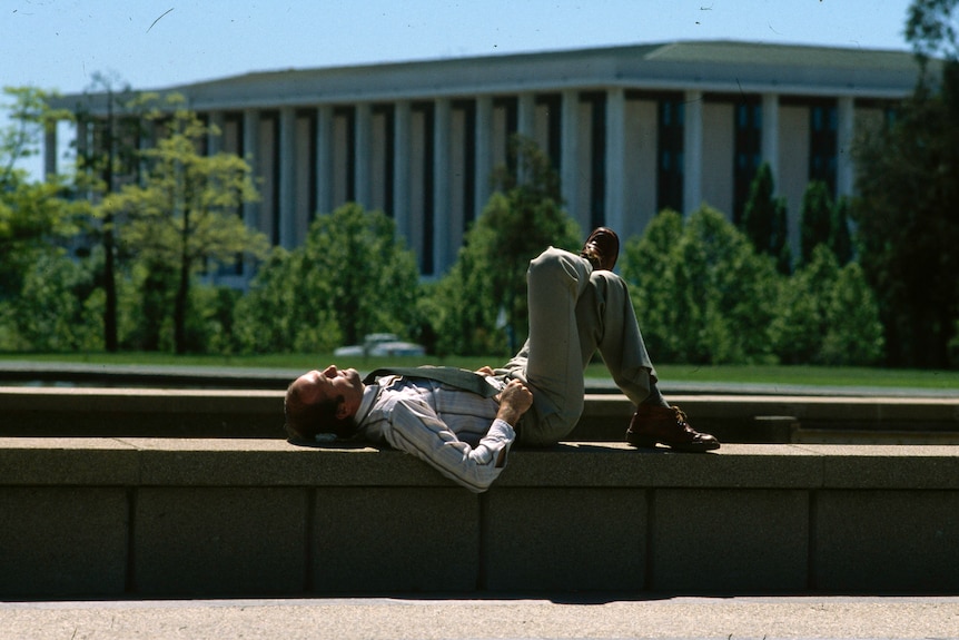 A man laying on a concrete bench with a grand, pillared building in the background.