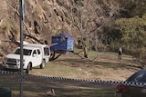 Police tape in front of an area of park at the base of cliffs. A blue tent can be seen, and a police officer with a tripod.