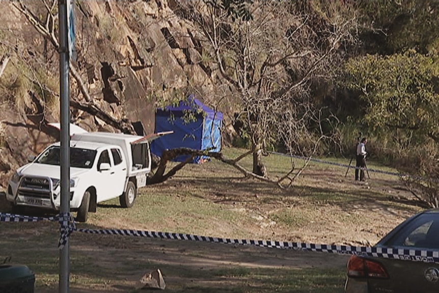 Police tape in front of an area of park at the base of cliffs. A blue tent can be seen, and a police officer with a tripod.