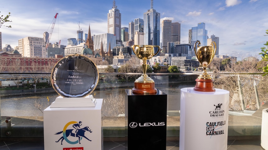 The Cox Plate, Melbourne Cup and Caulfield Cup trophies stand on pedestals with the Melbourne skyline in the background.