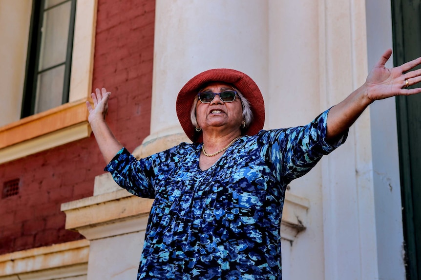 Woman wearing blue top, sunglasses and red hat with arms outstretched, stands in front of white and red brick building