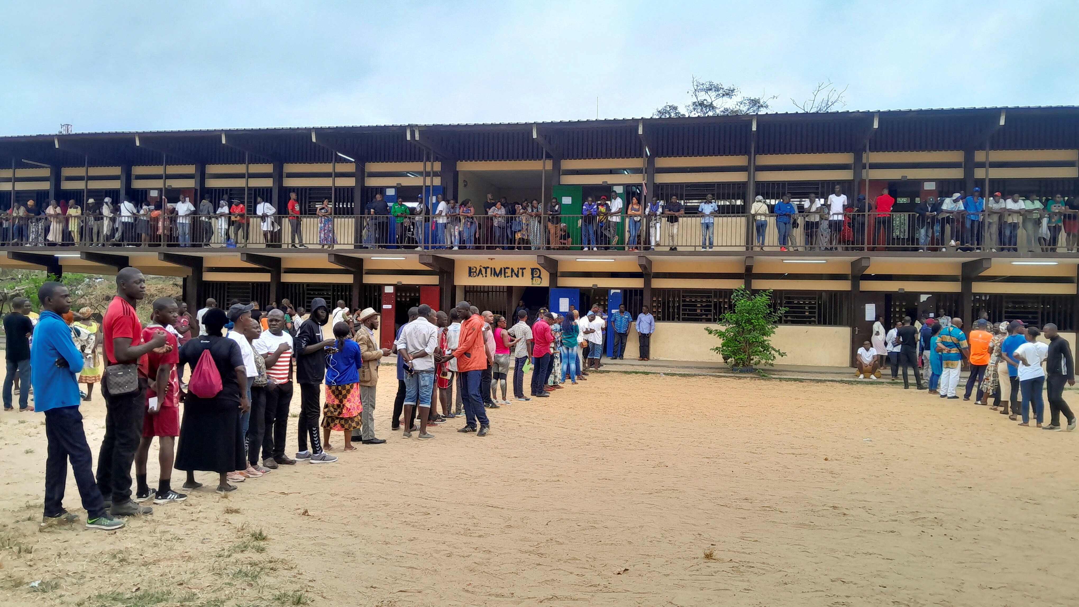 People standing in a line outside on sandy floor in front of a two storey bulding.