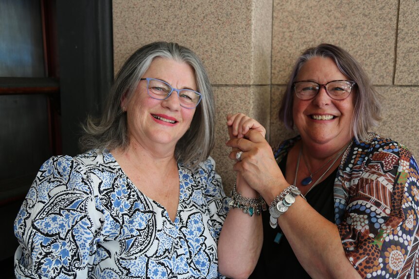 Two women both smiling widely and holding hands with raised arms to show the charm bracelets on their wrists.