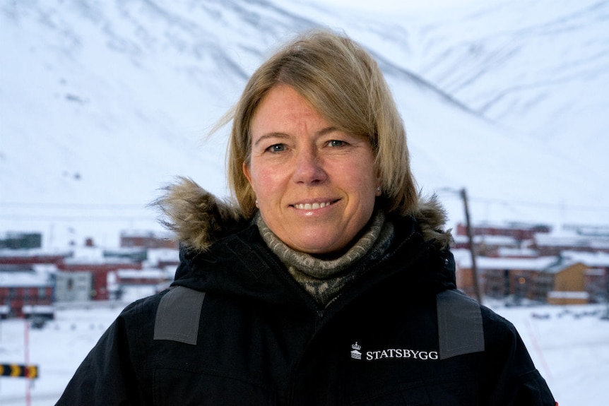 A blond woman wearing a ski jacket, standing outside in the snow, with a village in the background.