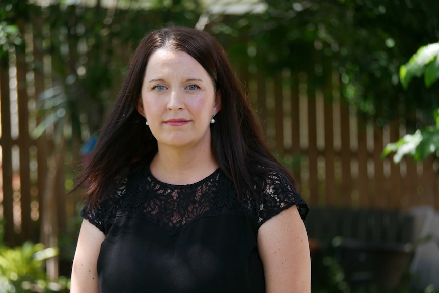 Angela Tallon stands outside her home, she is standing in front of a fence covered with foliage.