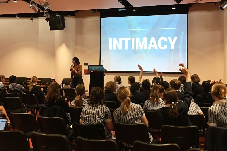 A woman holds a microphone in front of an image saying intimacy and a group of school children in chairs.