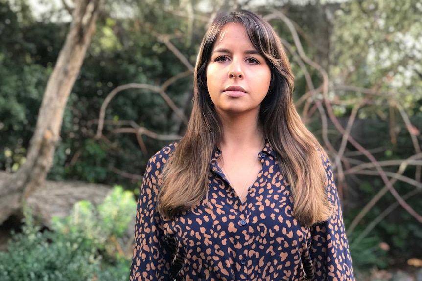 A young woman wearing a patterned blouse stands in front of a tree