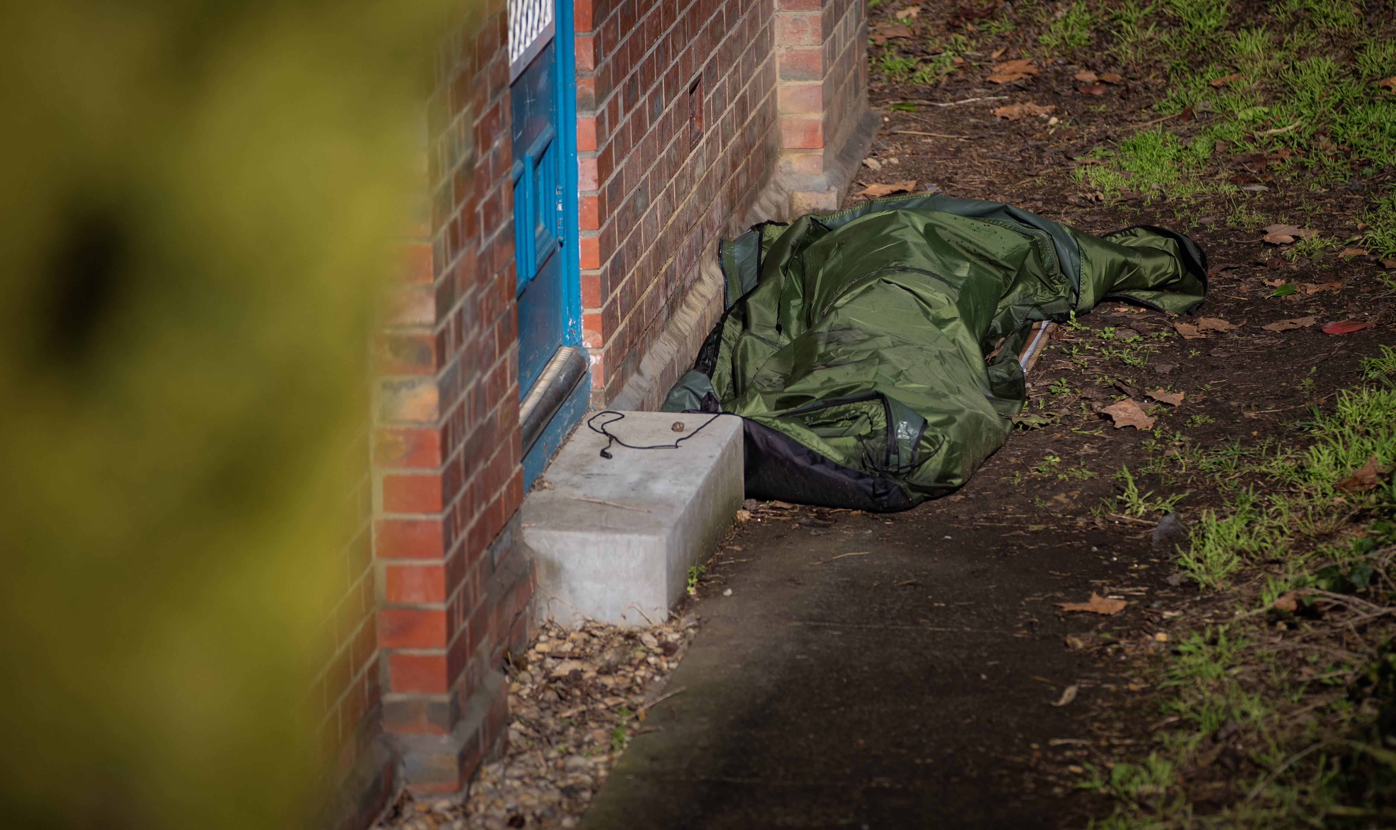 A person in a sleeping bag non identified sleeping outside against a brick wall.