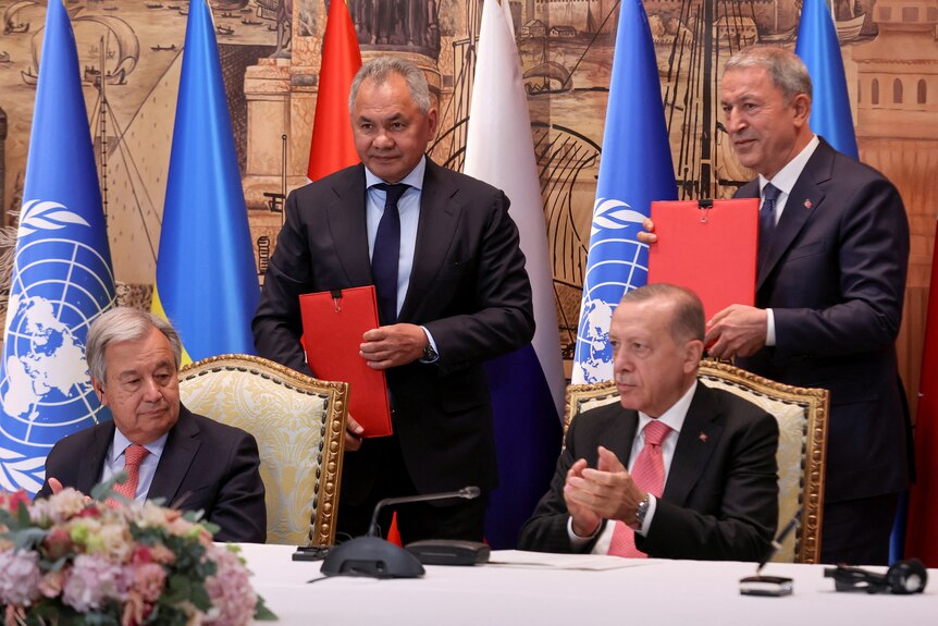 Four men in dark suits hold documents at an official function.