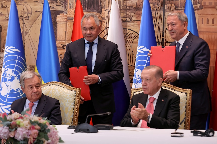 Four men in dark suits hold documents at an official function.