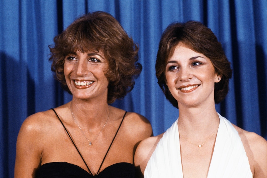 Two women smile together in front of a blue background. 