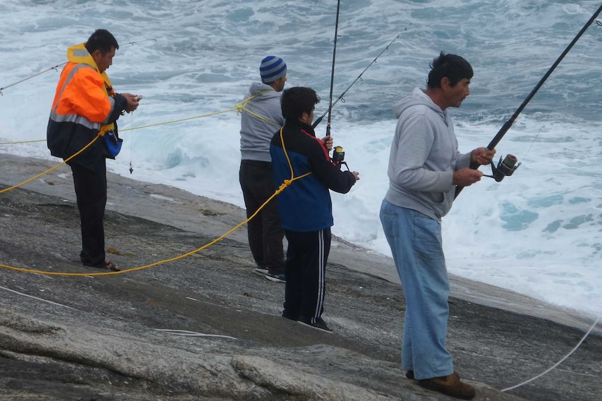Men tethered to lasso-style ropes fish on slanted rocks that slope down to the sea.