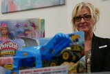 A blonde, bespectacled woman standing behind a pile of toys.