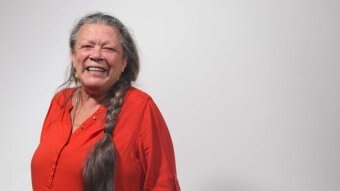 Woman with long plaited grey hair smiles at camera. She is wearing a red shirt, standing in front of a plain grey background.