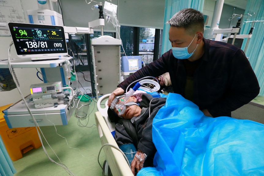 A man stands over a hospital bed where an elderly person is connected to several machines