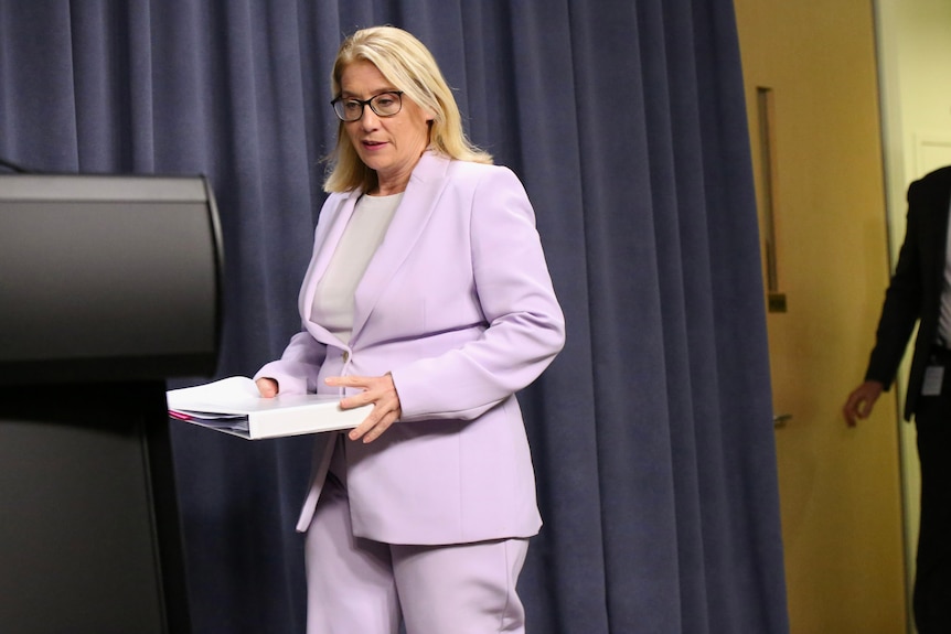 Rita Saffioti entre dans une salle pour une conférence de presse vêtue d'une veste et d'un pantalon lilas et d'un dossier blanc.